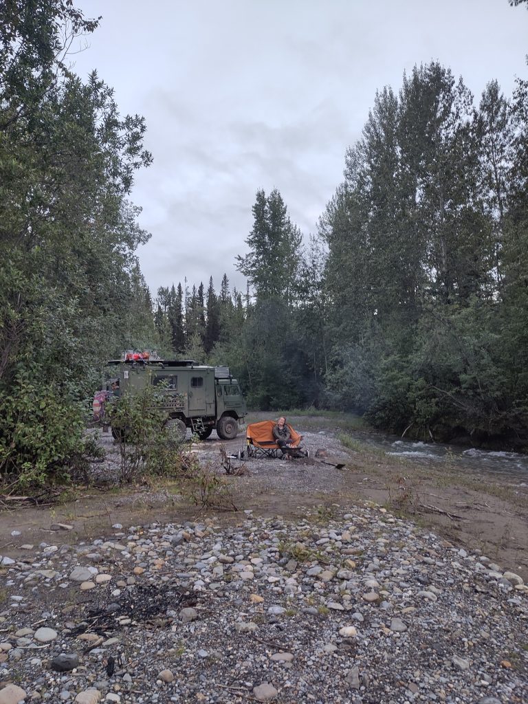 Bretti sitzt auf der campingbank vor dem Volvo an der feuerschale und neben ihr rauscht der Fluss