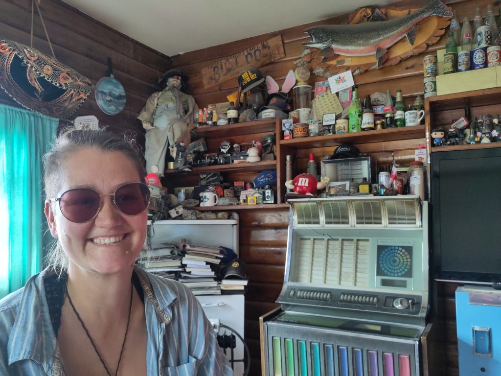 Foto: Bretti sitzt in der Snackbar, im Hintergrund viel Deko/Ramsch, von einem Sombrero bis zu einem Fisch, einer alten Jukebox? und viele Figuren