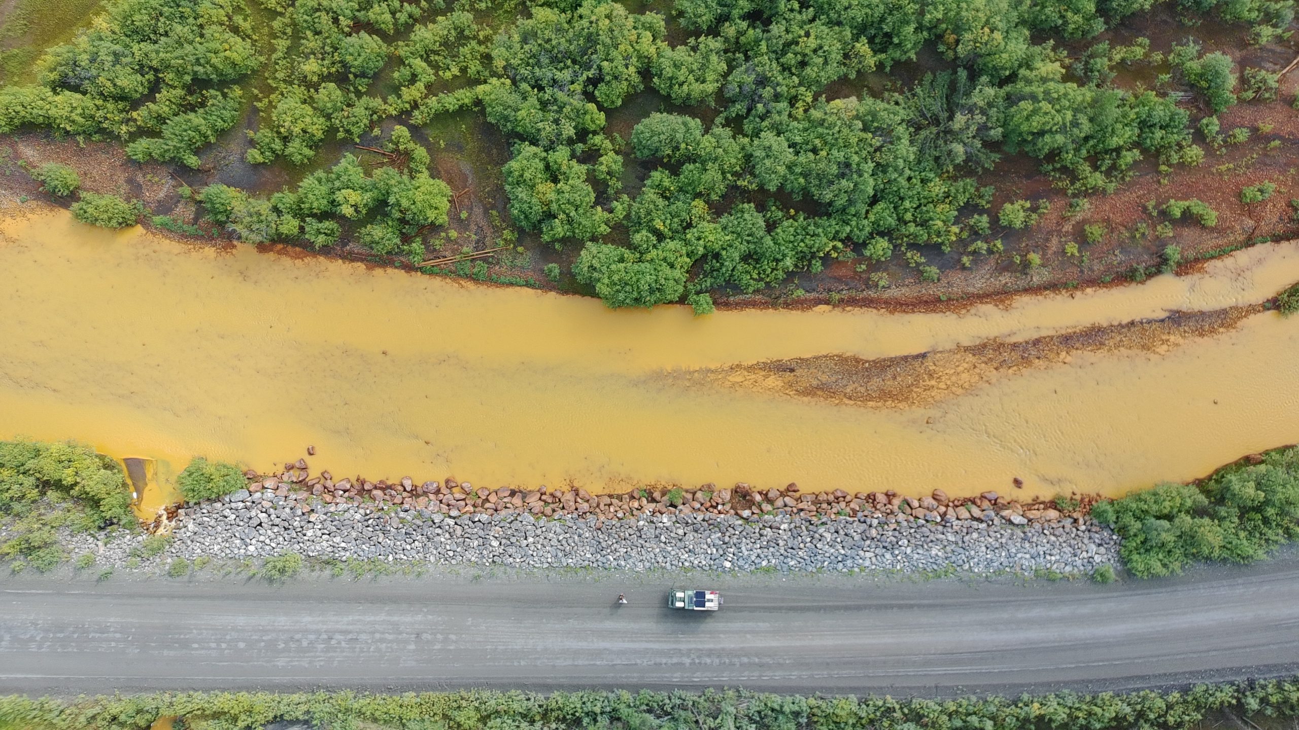 Foto: Drohnenaufnahme vom Volvo,auf dem grauen Dempster Highway, einem rostroten Fluss und grünem Wald